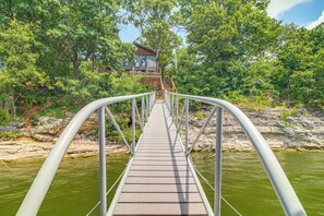A wooden bridge with metal railings leading to a building surrounded by trees and a body of water. - Private Hot Tub & Dock: Lakefront Grove Retreat! (Grove)