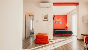 Una habitación de hotel moderna con suelo de madera, una pared de acento roja, una cama, un pequeño reposapiés naranja y un escritorio con una silla.