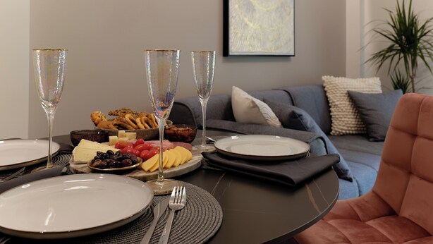 Une table à manger est dressée avec des assiettes, des couverts et une variété d’aliments, notamment du fromage, des olives et des fruits. On y trouve également deux flûtes à champagne.