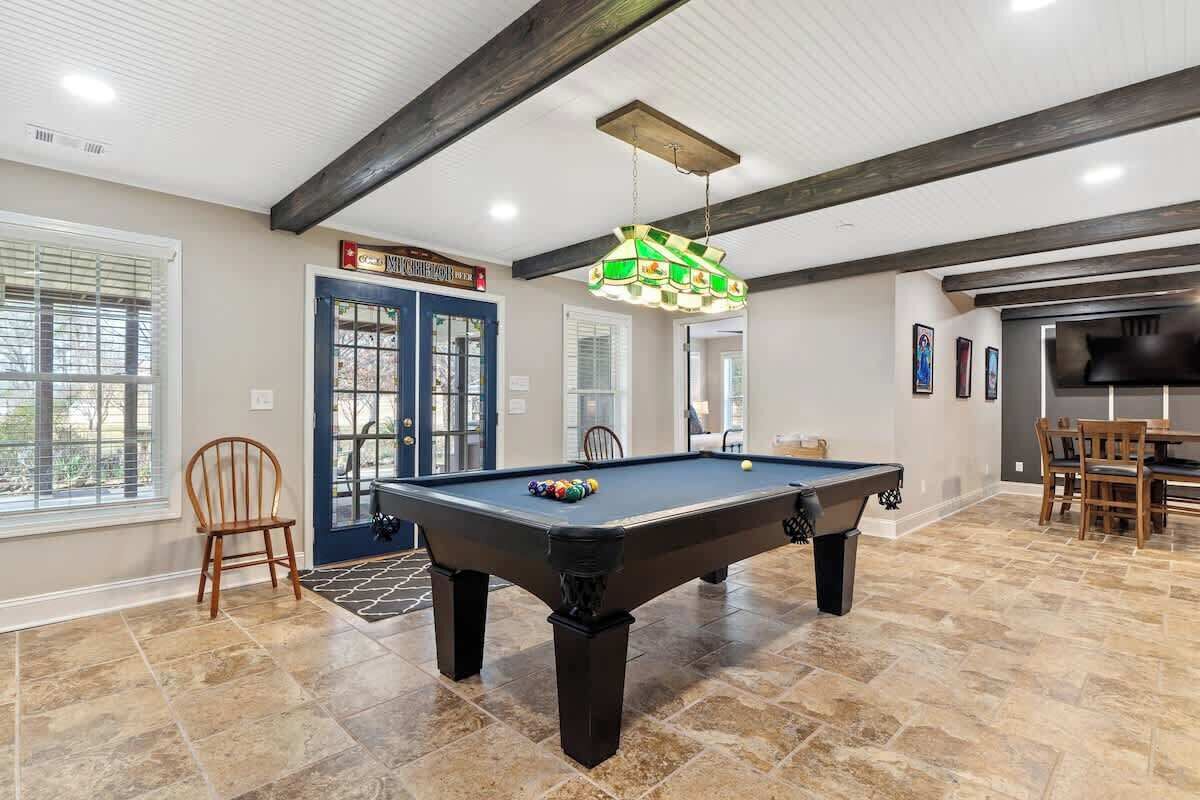 A pool table in a room with a high ceiling and exposed beams, a door labeled "Mansion Inn Hotel", and a dining area with chairs and a table.