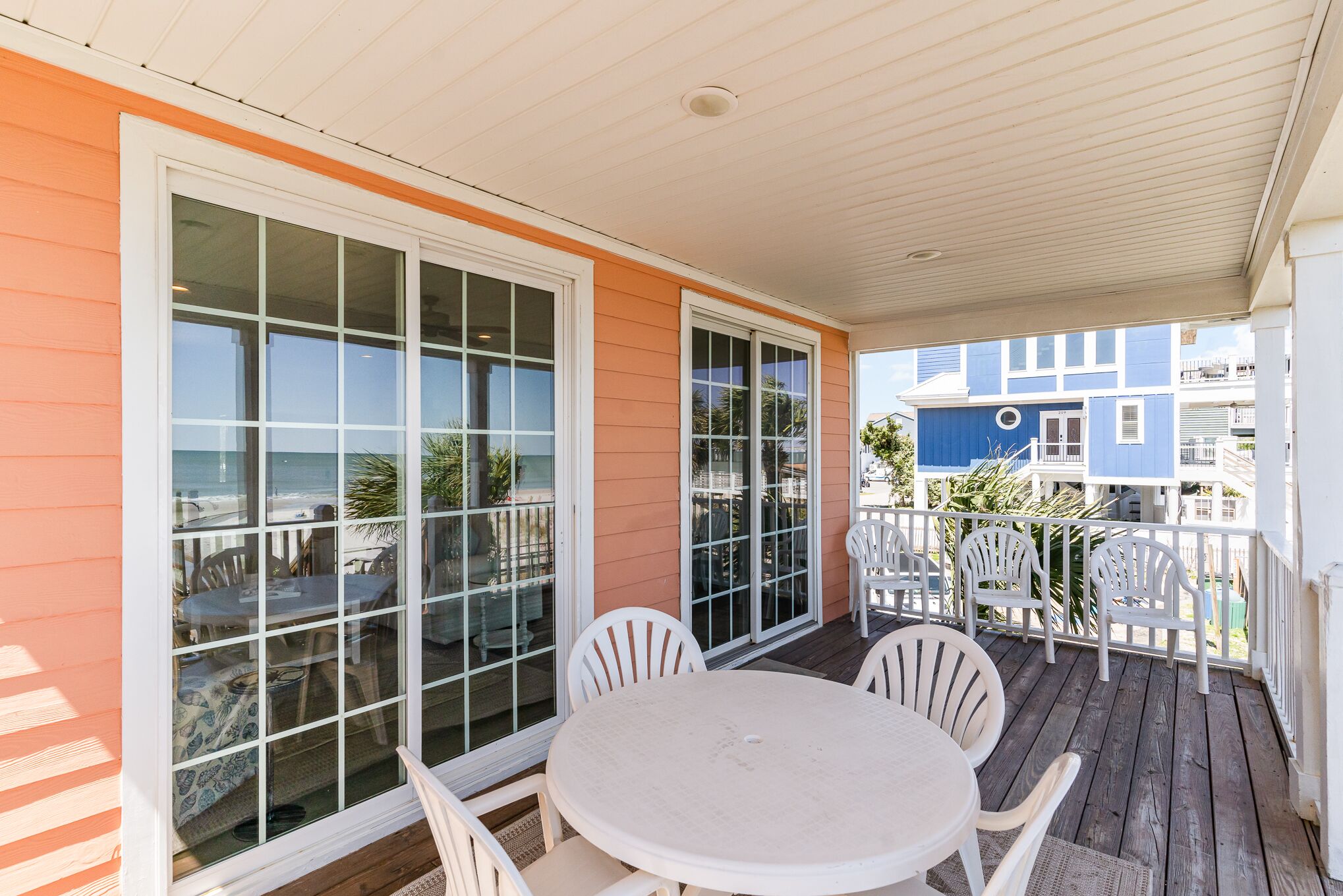 A covered outdoor patio with a round table and white chairs, large windows, and a view of the beach.