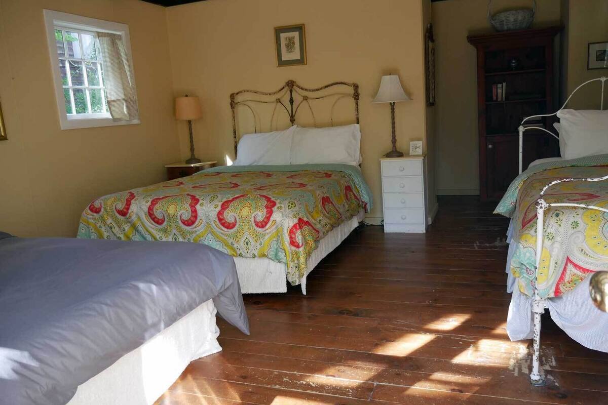 A room with two beds, a bedside table with a lamp, a wooden floor, and a window with blinds.