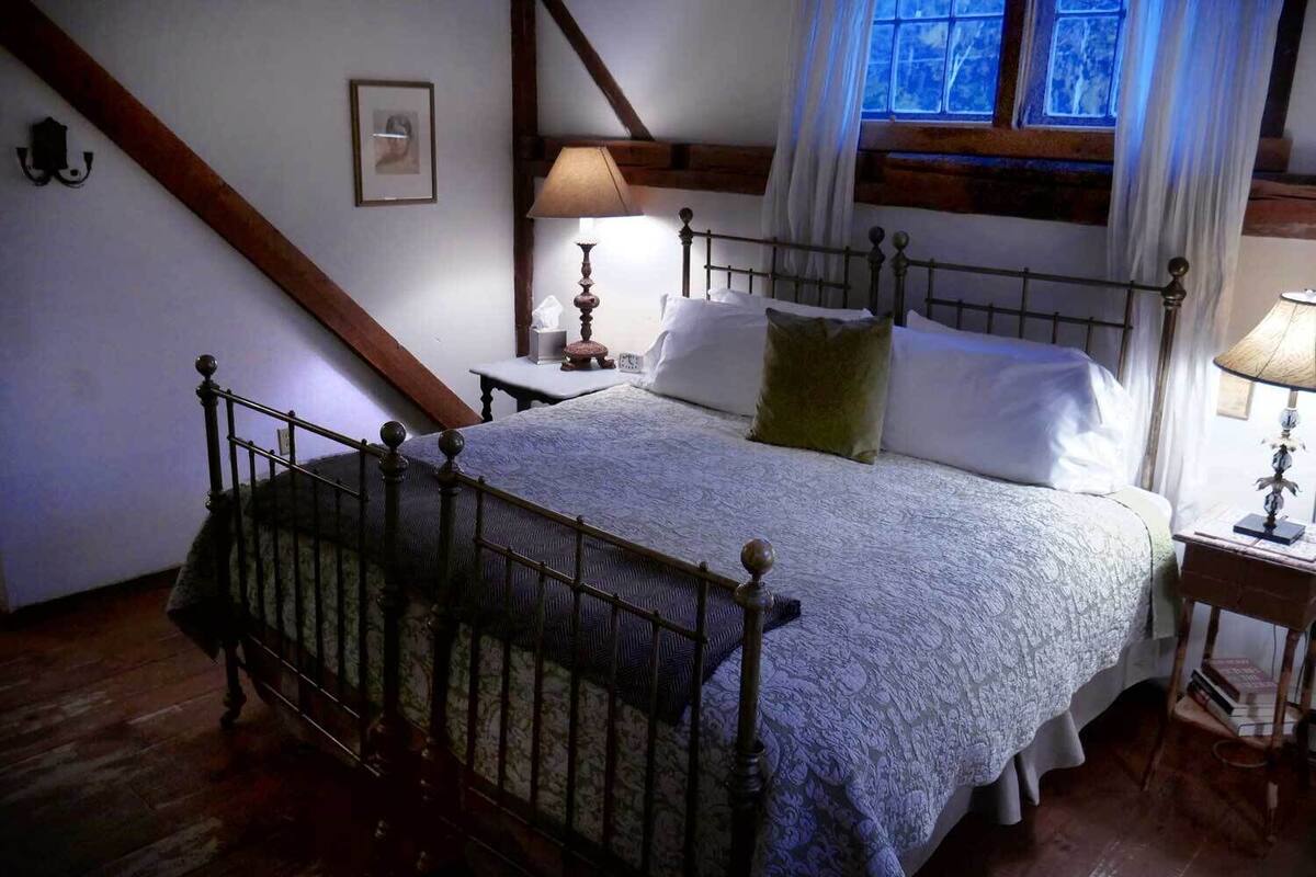 A bedroom with a four-poster bed, a nightstand, a lamp, a framed picture on the wall, and a window with curtains.