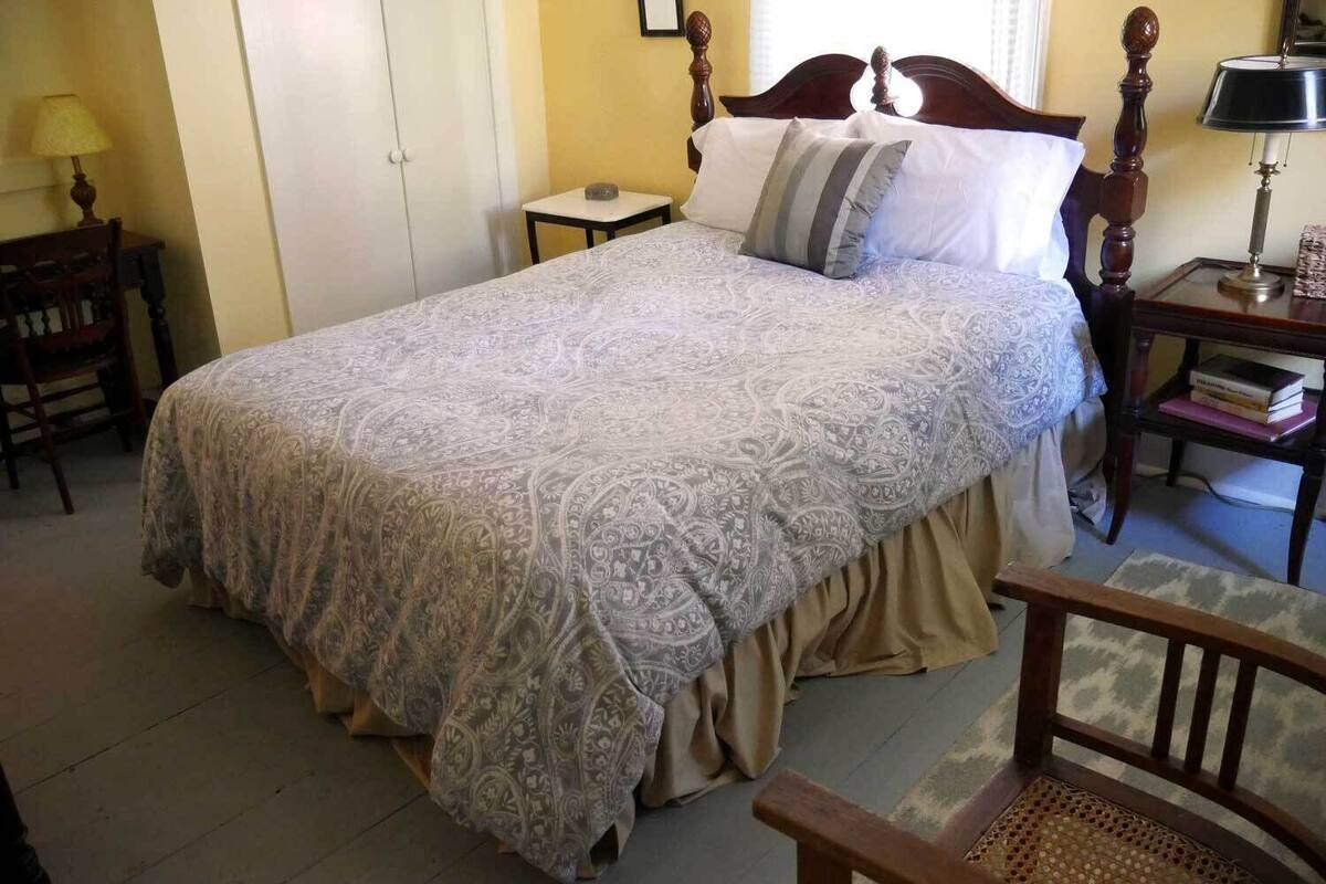 A neatly made bed with a patterned bedspread and pillows, a wooden nightstand with a lamp, and a chair in a room with a simple layout.