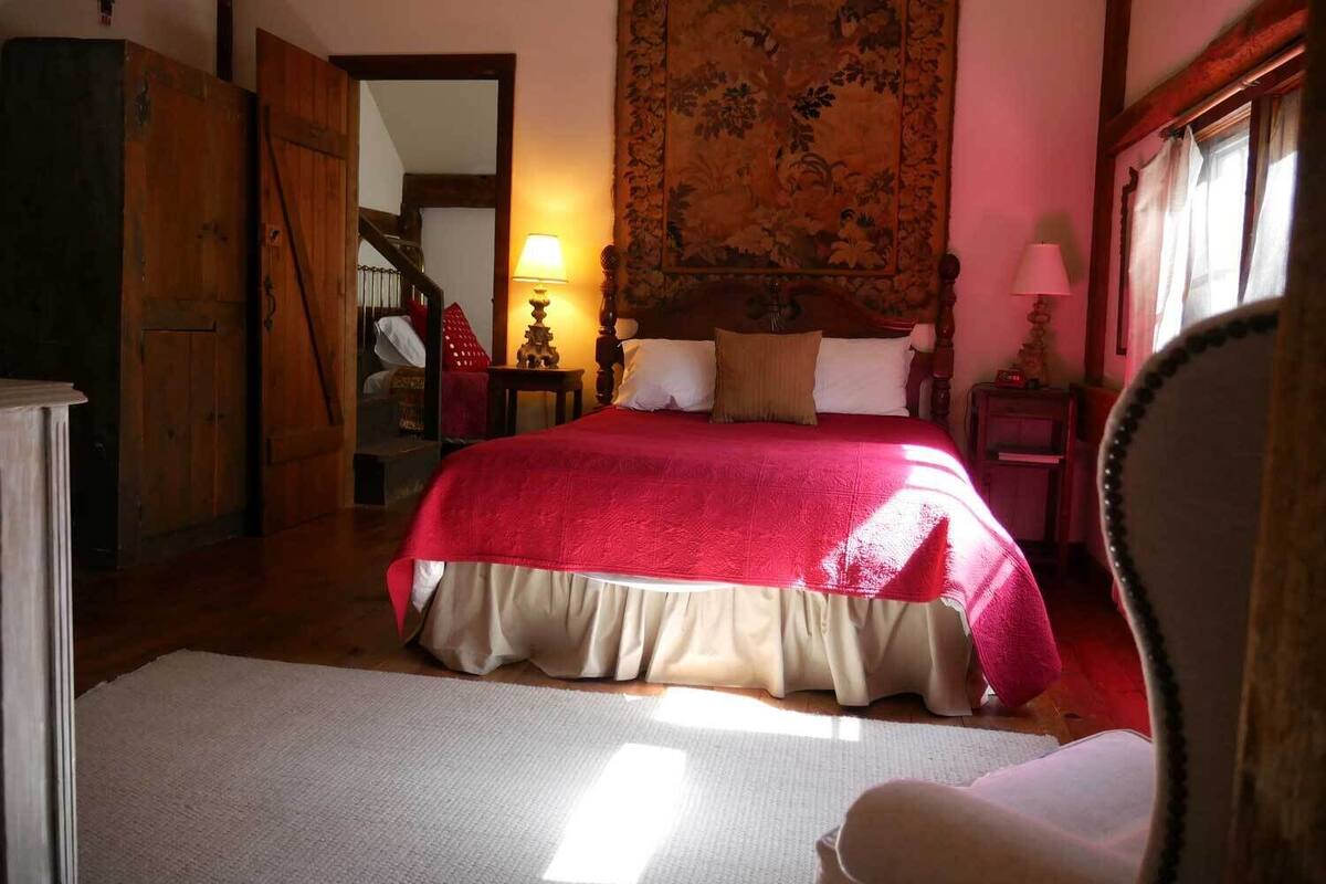 A bedroom with a large bed, a wooden wardrobe, a mirror, a lamp, and a tapestry on the wall.
