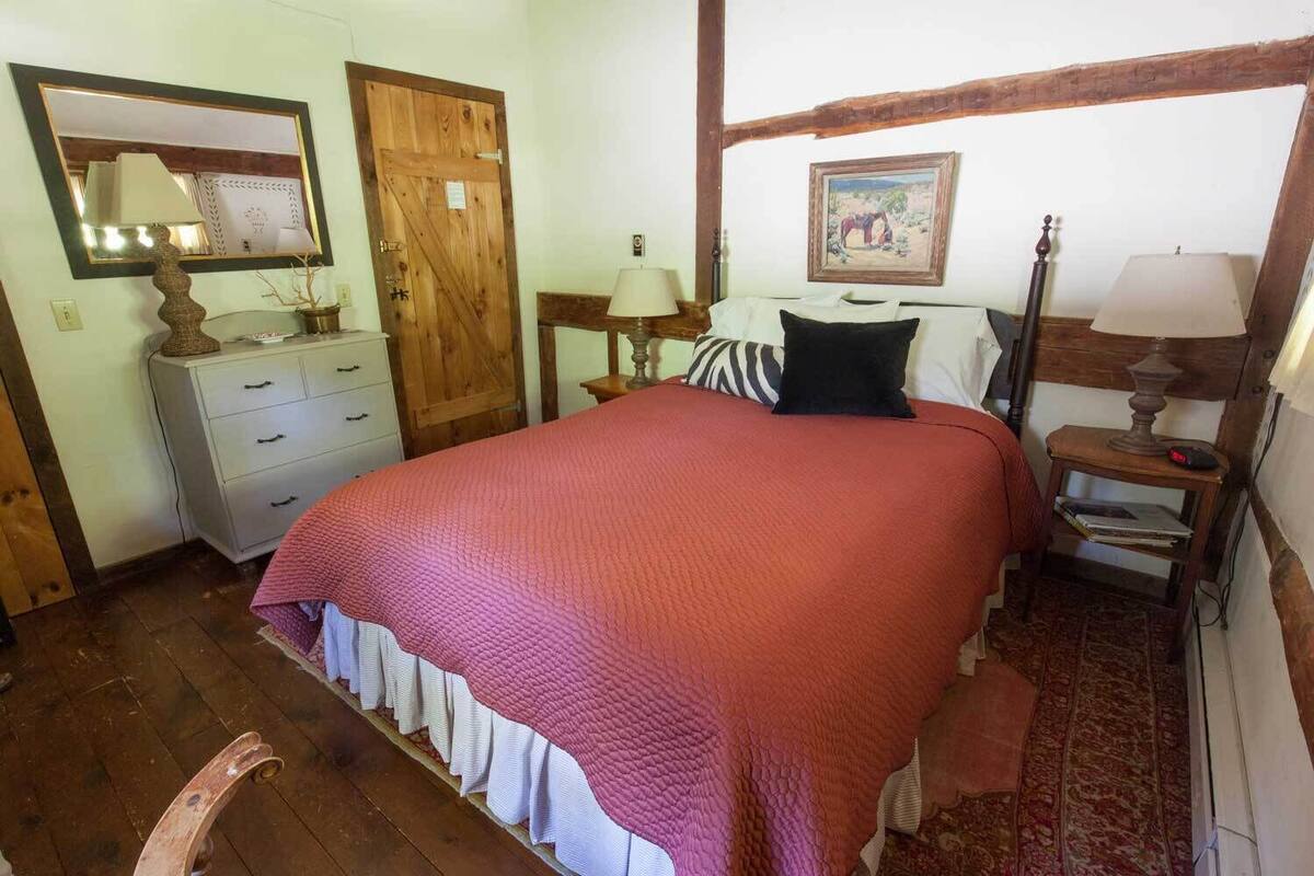 A bedroom with a large bed, a wooden wardrobe, a dresser, a nightstand, a lamp, and a painting on the wall.