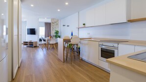 Una cocina moderna con armarios blancos, una encimera de madera y un horno empotrado. Hay una zona de comedor con una mesa y sillas, y un salón con sofá y televisor.
