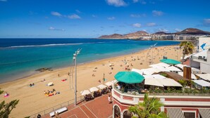 Una playa con cielo azul despejado, orilla arenosa y vista al mar. Hay sombrillas y gente en la playa.