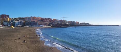 A beach with a sandy shore, a clear blue sea, and buildings in the background.