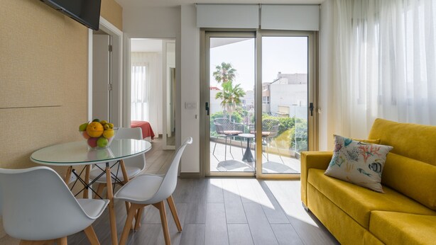 A modern living room with a yellow sofa, a glass dining table with chairs, and a balcony with a view of palm trees and buildings. - Triton By CanariasGetaway (Telde)