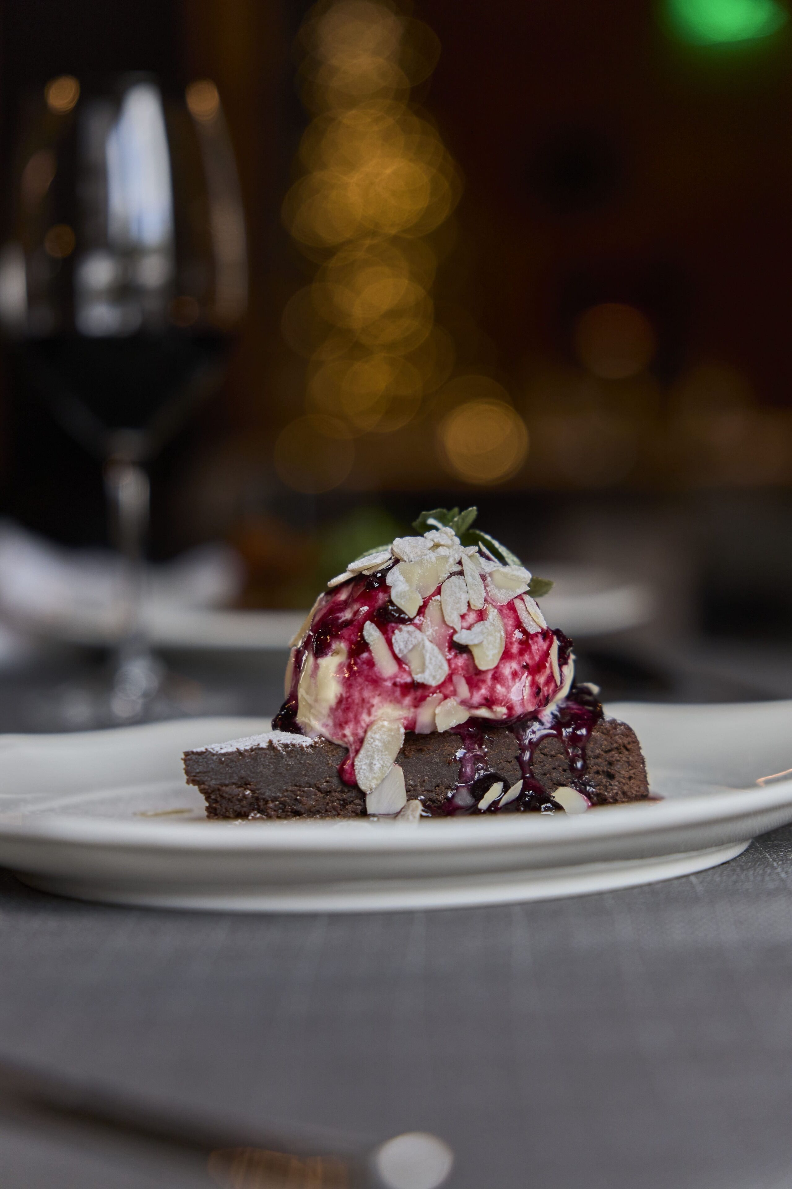 A dessert with a scoop of ice cream on top of a piece of cake, garnished with almonds and berries, served on a white plate.