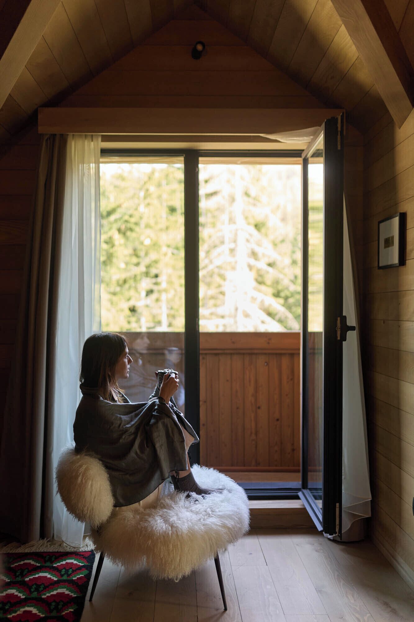 A person sitting on a fluffy chair by a large window with a view of trees.