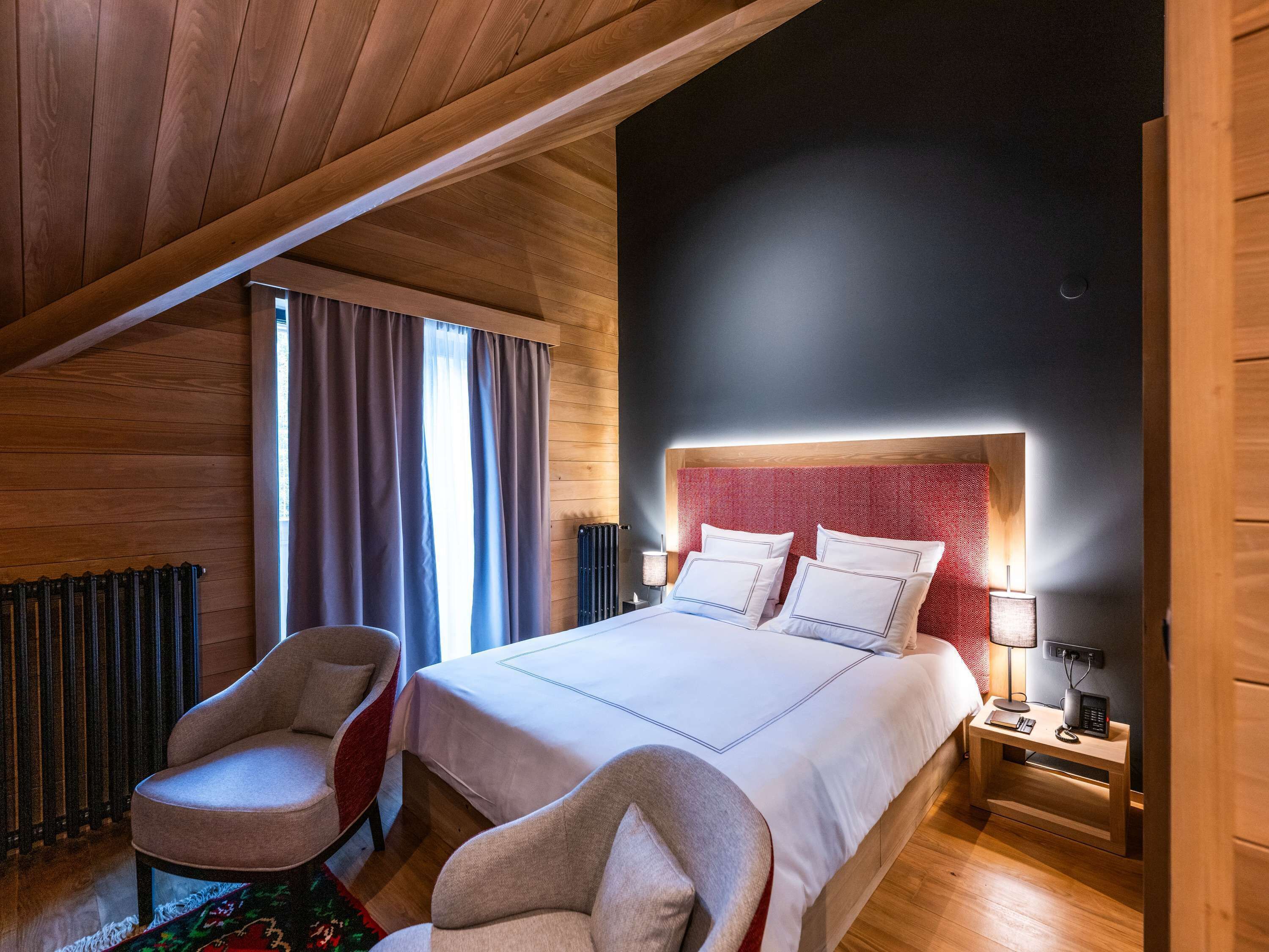 A bedroom with a wooden ceiling, a bed with white bedding, two bedside tables with lamps, and a chair.