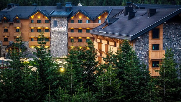 A multi-story hotel with a stone facade and wooden balconies, surrounded by dense forest.