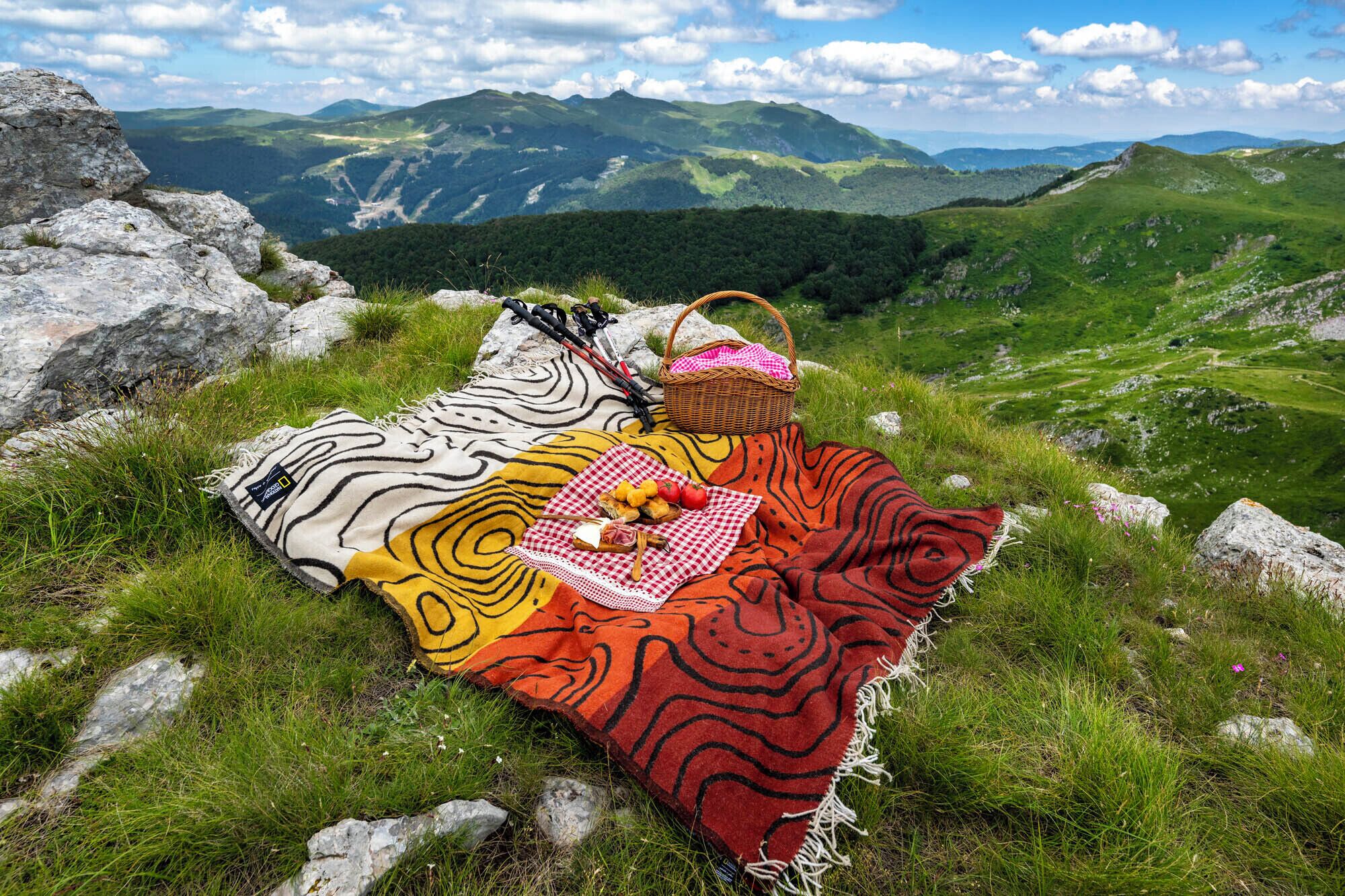 A picnic setup on a grassy hill with a blanket, a basket, and food items.