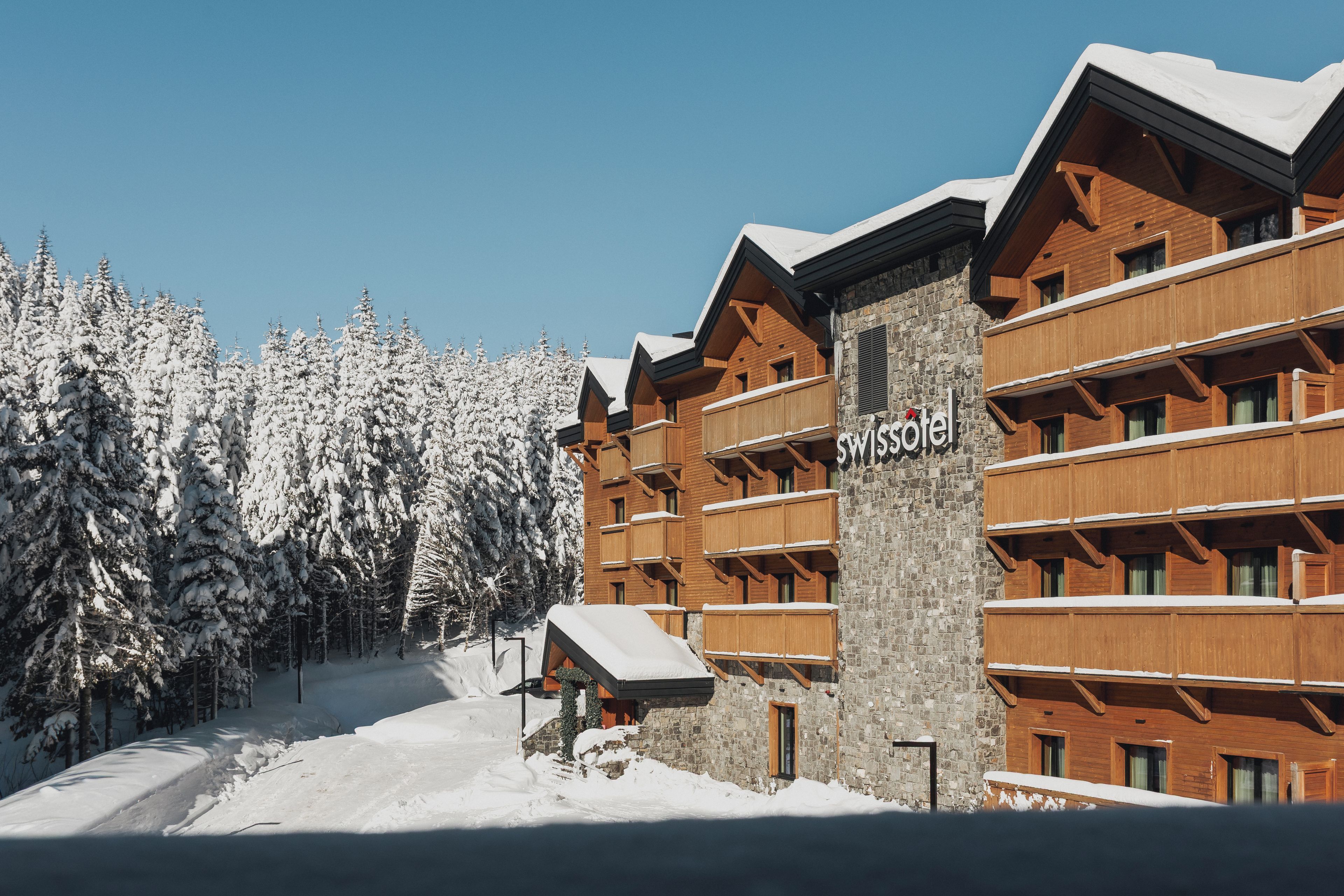 A hotel with a stone facade and wooden balconies, surrounded by snow-covered trees.