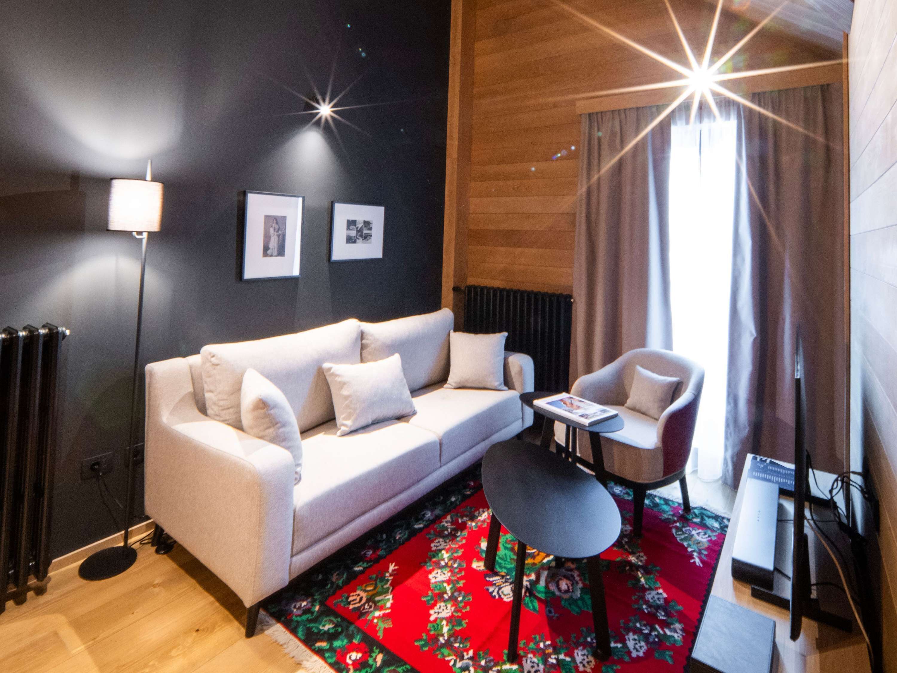 A modern living room with a sofa, armchair, and coffee table. There are framed pictures on the wall, a floor lamp, and a radiator.