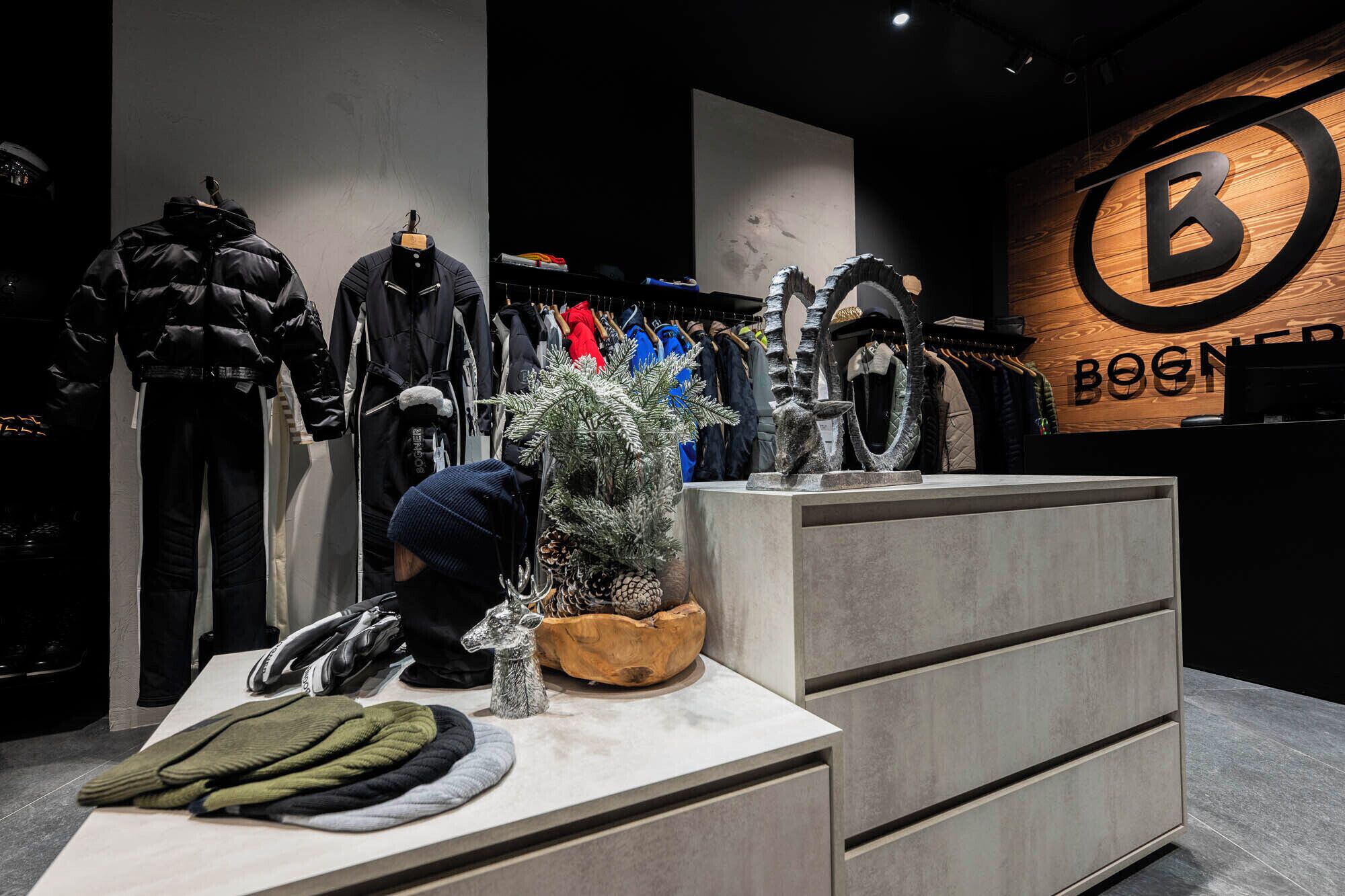 A clothing store with racks of jackets and coats, a display table with hats and scarves, and a wooden sign with the store's name.