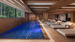 A modern indoor swimming pool with wooden beams, lounge chairs, and a view of the forest.