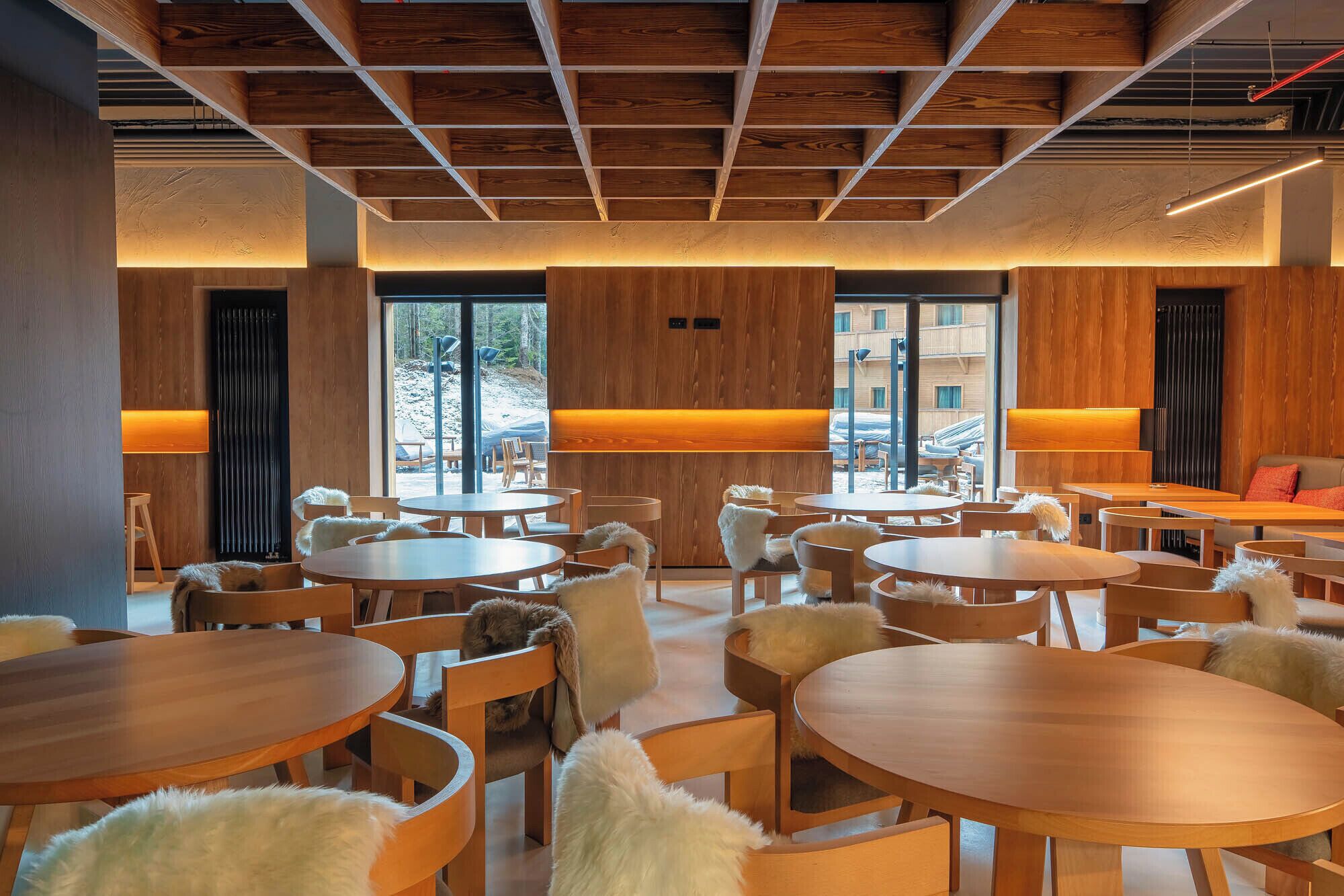 Un interior moderno con mesas y sillas redondas, paneles de madera y ventanas amplias que ofrecen vistas al exterior.