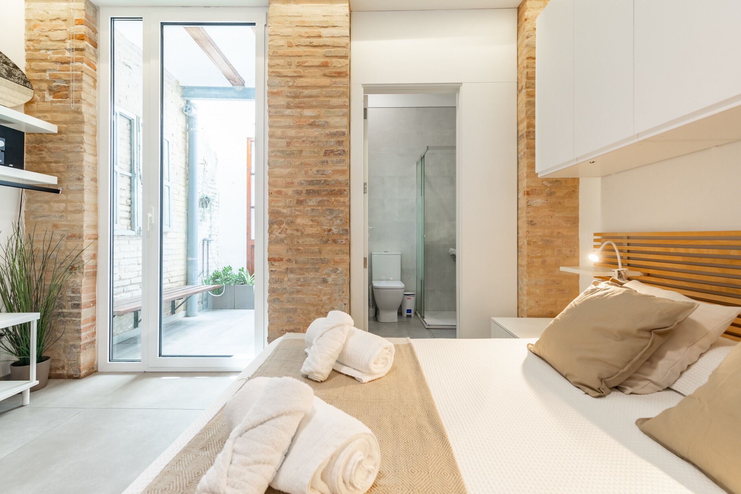 Quarto moderno com cama, mesa de cabeceira, um armário para objetos pessoais, janela e banheiro visível por uma porta aberta.