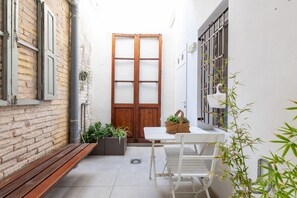 A narrow outdoor corridor with a wooden door, a white table with chairs, a bench, and potted plants.