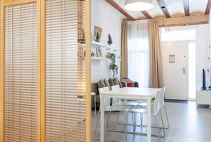 Un salon moderne avec une table à manger blanche, un canapé avec des coussins et une cloison en bois avec des stores.