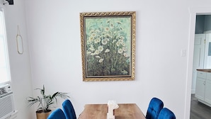 Un coin repas avec une table en bois, des chaises à l’assise bleue, un tableau encadré accroché au mur, une plante en pot et un mur blanc.