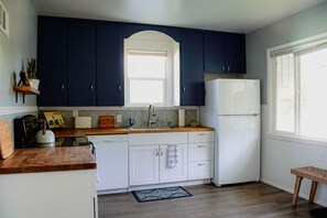 Cocina con armarios azul oscuro, electrodomésticos blancos, encimera de madera y ventana que proporciona luz natural.