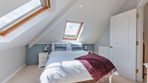 Une chambre avec une lucarne, un lit recouvert d’une couverture blanche et rose, et une table de chevet avec une lampe.