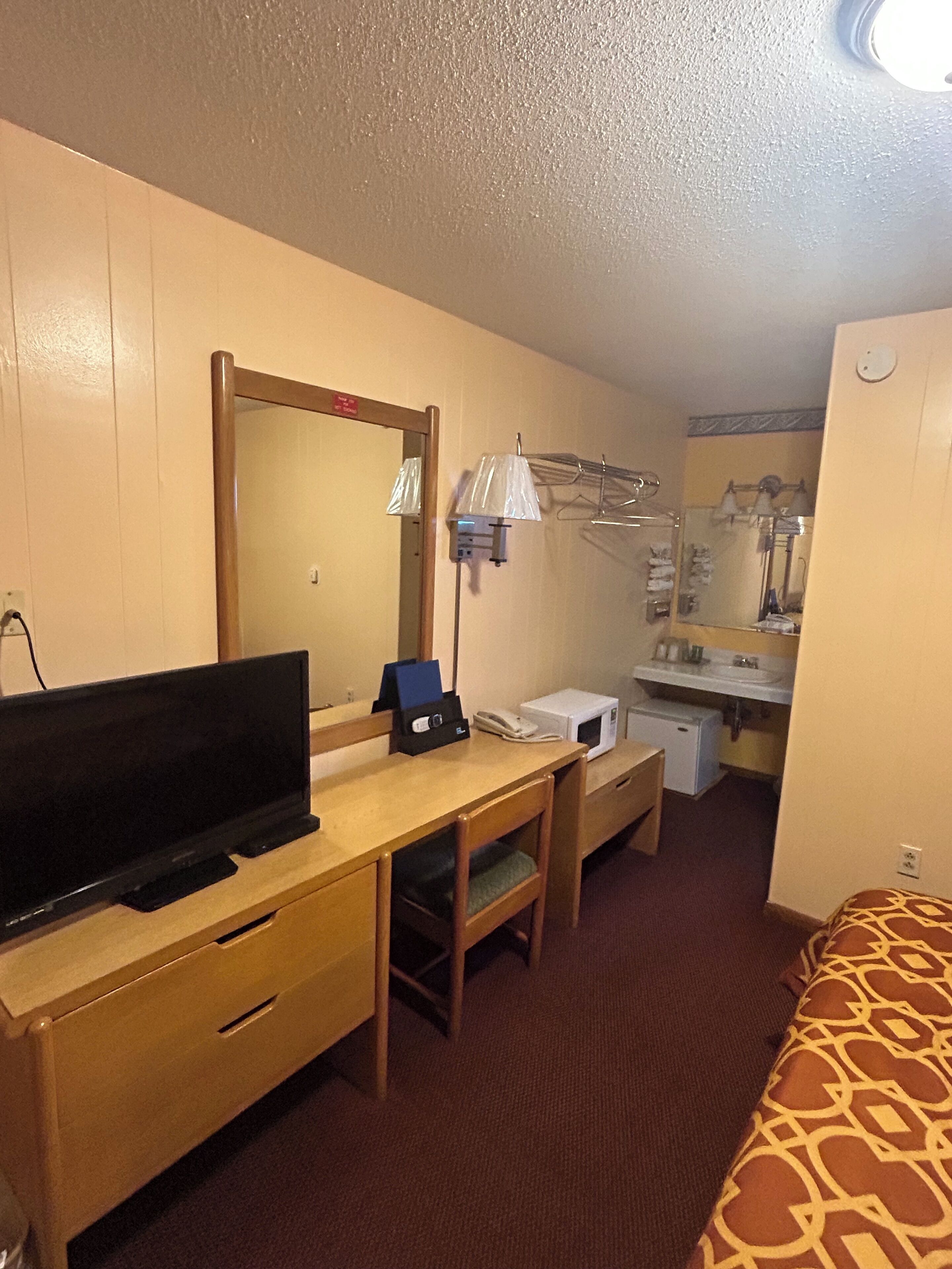 A hotel room with a bed, a desk with a television, a telephone, a microwave, and a small bathroom with a sink and mirror.
