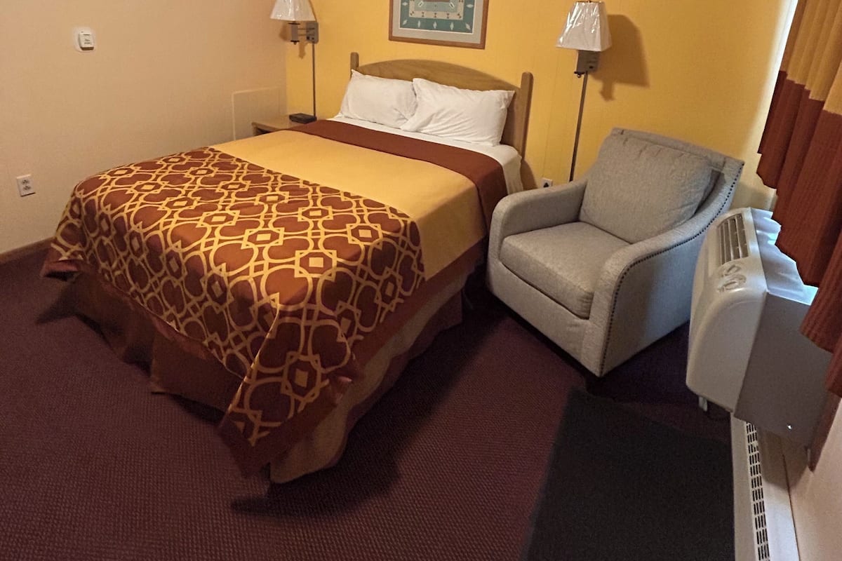 A hotel room with a bed, a chair, a heater, and a wall-mounted lamp.