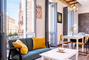 A modern living room with a sofa, yellow cushions, a white table, and a city view through large windows.