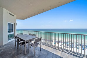 Een balkon met een tafel en stoelen, met uitzicht op een strand met parasols en mensen.