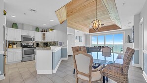 A modern kitchen with white cabinets, stainless steel appliances, and a large island. There is a dining area with a glass table and wicker chairs. - Stunning Beach, Golf Course and Bay Views!  Empress 1103 (Miramar Beach)