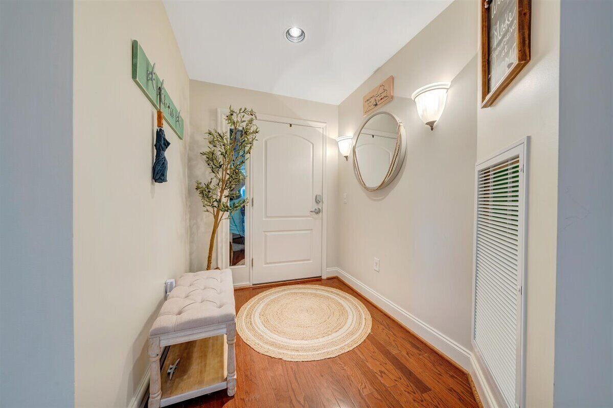 A hallway with a white door, a round woven rug, a bench, a wall-mounted mirror, and a wall-mounted light fixture.