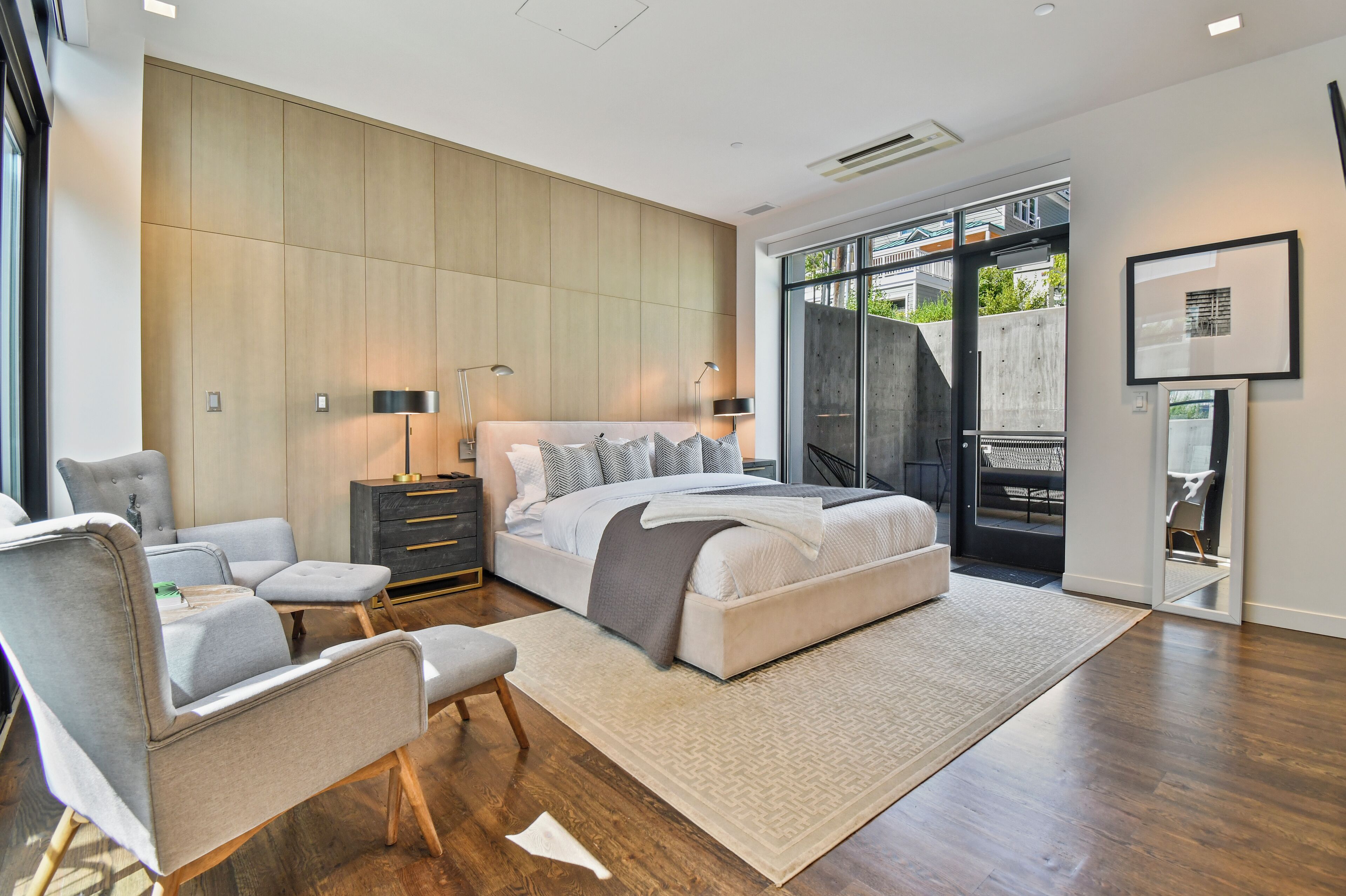 Un dormitorio moderno con una cama grande, sillas tapizadas en gris, una mesita de noche oscura y un amplio ventanal con vistas al exterior.