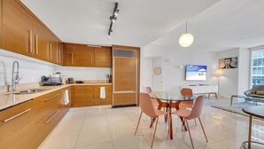 A modern kitchen with wooden cabinets, a dining area with a glass table and pink chairs, and a living area with a sofa and TV. - Icon Luxury 34th Floor Amazing Oceanview, Brickell (Miami)