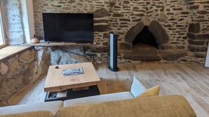 A stone fireplace with a flat-screen TV mounted above it, a wooden coffee table with a magazine, and a sofa in a room with wooden flooring. - Gîte Atypique Avec Services Tout Inclus, à L'orée D'une Forêt Privée (Le Mené)