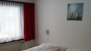 Un lit simple avec une literie à motifs floraux, dans une pièce dotée d’une fenêtre, de rideaux rouges et d’un petit luminaire fixé au mur.