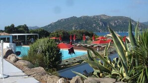 Zona de piscina con sombrillas rojas, vista despejada al mar y montañas al fondo.