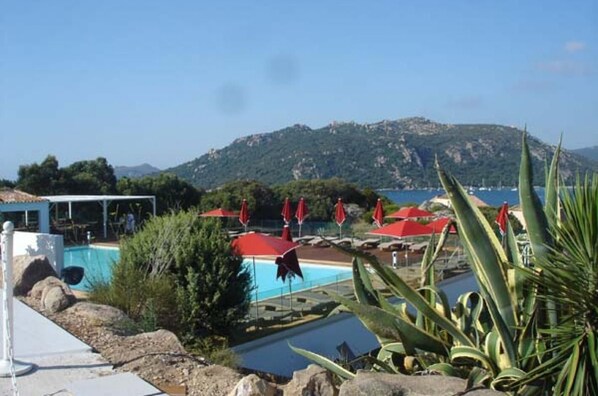 A pool area with red umbrellas, a clear view of the sea, and a mountainous backdrop.