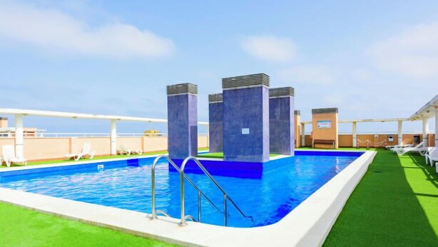 A rooftop swimming pool with a clear blue pool, surrounded by green artificial grass, and white lounge chairs.