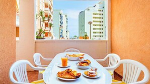 A dining area with a table set for two, featuring a meal with pizza and a glass of orange juice. The room has a large window offering a view of tall buildings and palm trees.
