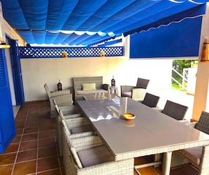 Un área de comedor al aire libre con mobiliario de mimbre, una mesa y toldos azules.