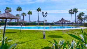 A swimming pool surrounded by grass, palm trees, and a thatched-roof structure. - Beachfront 3 Bed Apt San Luis De Sabinillas (San Luis de Sabanillas)