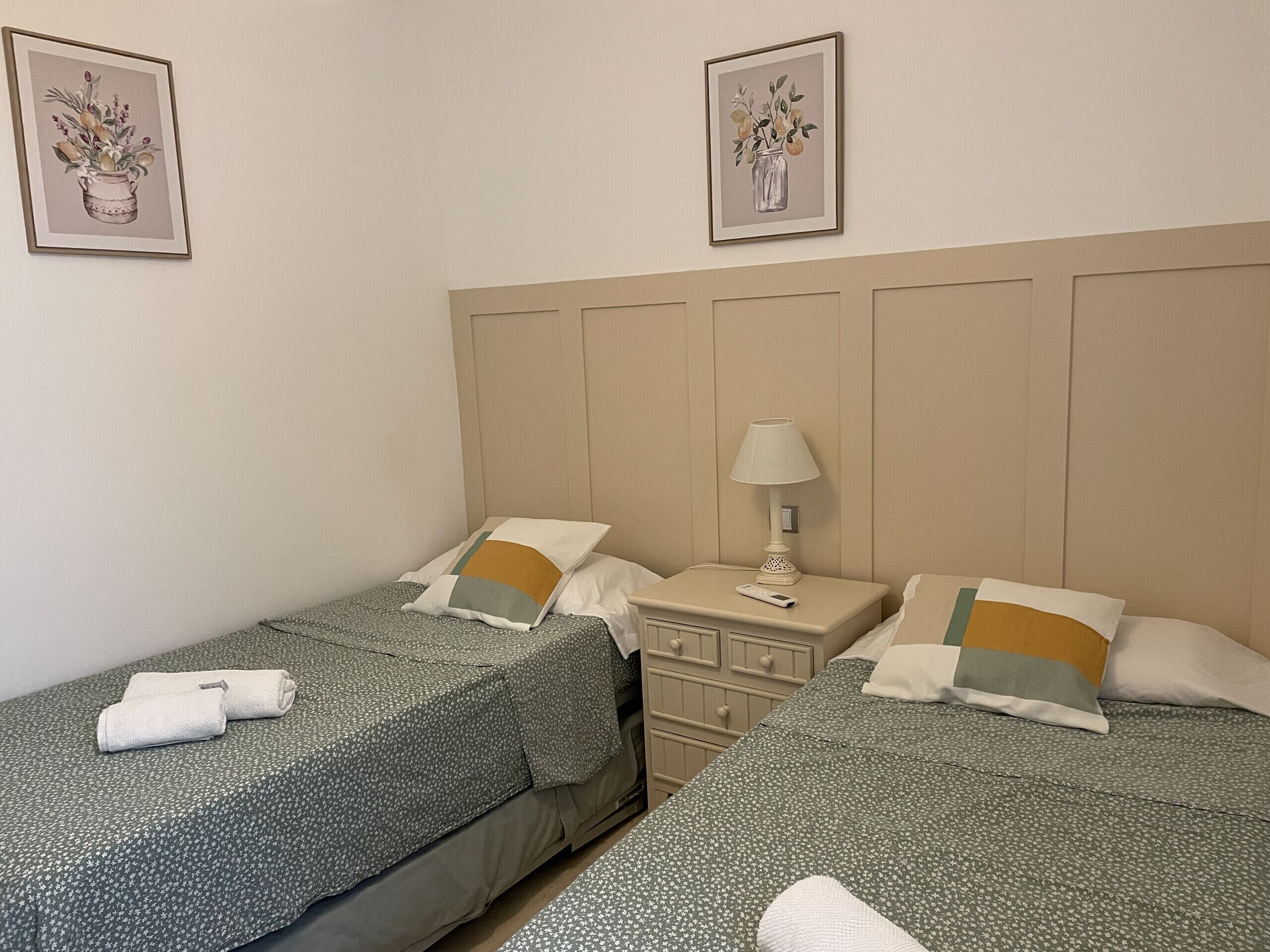 A hotel room with two single beds, a nightstand with a lamp, a dresser, and two framed floral artworks on the wall.