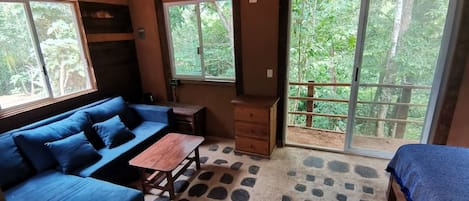 A room with a sofa, a table, and a bed. There are windows and a view of greenery outside.
