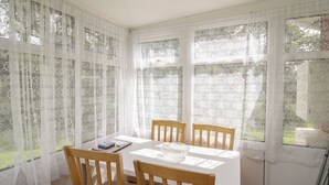 A dining area with a table and four wooden chairs, large windows with lace curtains, and a view of greenery outside. - Chalet 64 (Tywyn)