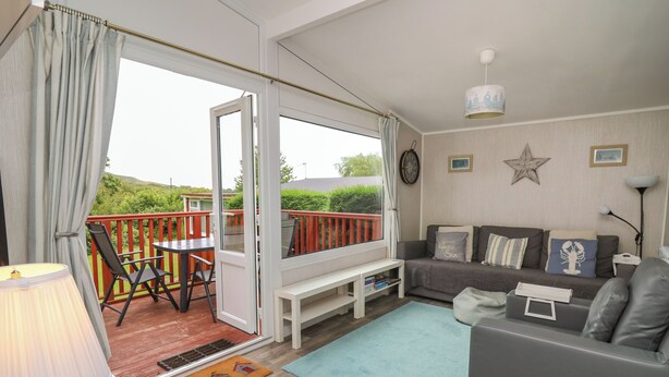 A cozy living room with a sofa, a coffee table, and a dining area with a table and chairs. There is a balcony with a railing and a view of greenery outside. - Chalet 64 (Tywyn)